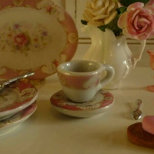 May include: Miniature porcelain tea set with pink floral accents. Includes a teacup, saucer, plates, and a serving platter. A small white vase holds pink and yellow roses. A pink candle holder and a pastry are also present.