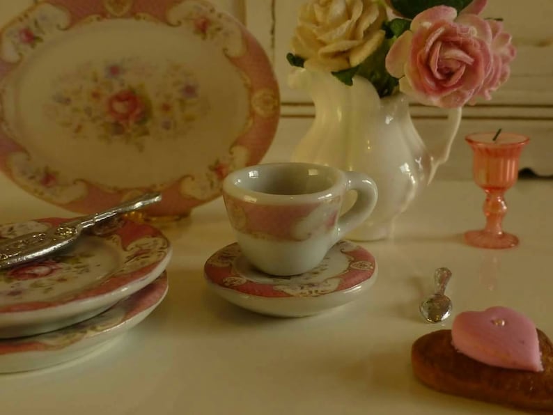 May include: Miniature porcelain tea set with pink floral accents. Includes a teacup, saucer, plates, and a serving platter. A small white vase holds pink and yellow roses. A pink candle holder and a pastry are also present.