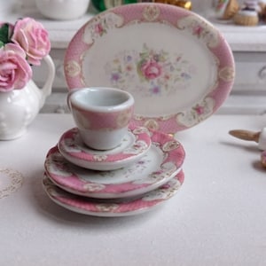 May include: A miniature porcelain tea set with a pink floral design. The set includes a teacup, saucers, and a serving plate. The edges of the pieces are trimmed with pink. The set is part of a dollhouse collection.