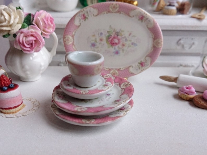 May include: A miniature porcelain tea set with a pink floral design. The set includes a teacup, saucers, and a serving plate. The edges of the pieces are trimmed with pink. The set is part of a dollhouse collection.
