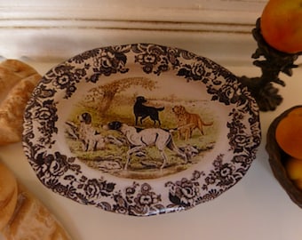 May include: A white oval ceramic dish with a black and white floral pattern around the edge. The center of the dish features a hunting scene with three dogs, a black and white dog, a brown and white dog, and a white dog with brown spots. The scene is set in a forest with trees and bushes.