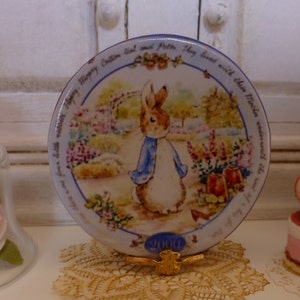 May include: A blue and white ceramic plate with a picture of Peter Rabbit walking through a garden. The plate has a blue border with white text that reads "Mary Caden bal and Patte. They lived with thein other worth the wo" and the number "200070".