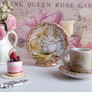 Könnte beinhalten: Eine weiße Teetasse und Untertasse mit einem Blumenmuster und einem Hasendesign. Die Teetasse steht auf einer weißen Untertasse mit goldener Verzierung. Die Teetasse und Untertasse stehen auf einem weißen Tisch mit einem rosa-weißen Blumenhintergrund. Der Text "Josephine Queen Rose Garden" ist im Hintergrund sichtbar.