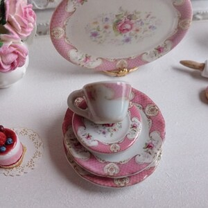 May include: Miniature pink and white porcelain tea set with floral designs. The set includes a teacup, plates, and a serving platter. A small cake and a vase of pink roses are also present, creating a charming, delicate scene.