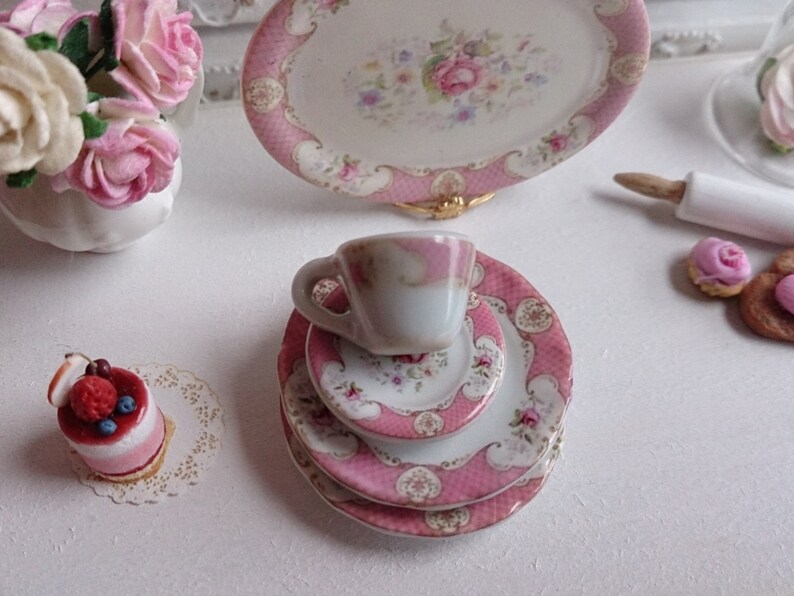 May include: Miniature pink and white porcelain tea set with floral designs. The set includes a teacup, plates, and a serving platter. A small cake and a vase of pink roses are also present, creating a charming, delicate scene.