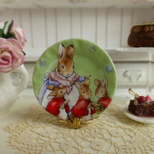 Peut inclure: Une assiette en céramique verte et blanche avec une image d'une mère lapin et de trois lapins. L'assiette est sur un support doré et une dentelle blanche.
