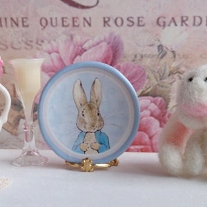 Puede incluir: Un plato de cerámica azul y blanco con una imagen de Peter Rabbit. El plato está en un soporte dorado.