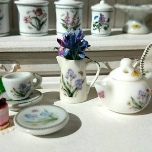 Könnte beinhalten: Ein Miniatur-Tee-Set mit Teekanne, Milchkännchen, Zuckerdose und Tasse mit Untertasse. Das Set ist mit floralen Mustern in Blau-, Lila- und Grüntönen verziert. Das Set wird auf einer weißen Oberfläche mit einem kleinen Kuchen und einer Flasche Champagner präsentiert.