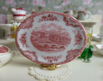 May include: A miniature, oval-shaped plate with a red and white floral and landscape design. The plate is held in a gold-colored stand and is part of a miniature tea set. The plate has a scalloped edge.