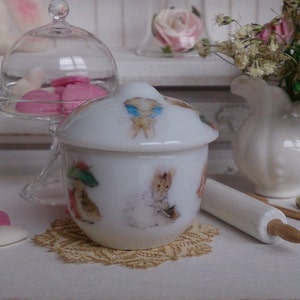 Puede incluir: Tarro de cerámica blanca con tapa, con un diseño caprichoso de conejos y flores. El tarro está sentado en un encaje y tiene un rodillo de amasar blanco al lado.
