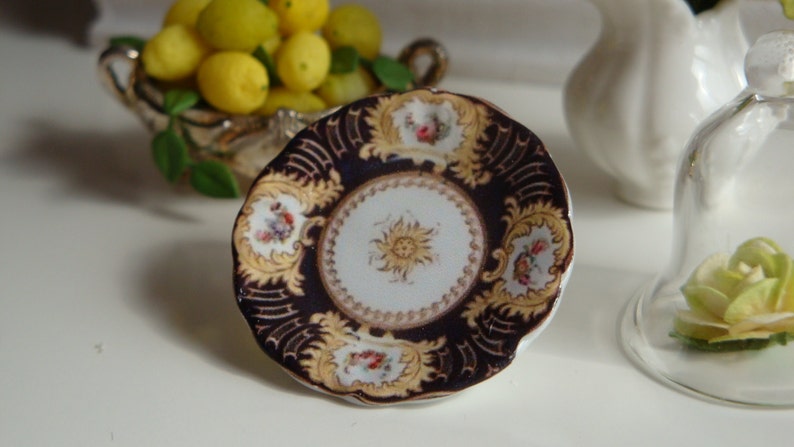May include: A small, decorative plate with a dark blue center and gold accents. The plate has a white center with a floral design and a gold rim.