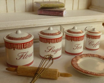 May include: Four white ceramic canisters with red and white gingham trim and black lettering. The canisters are labeled "Flour", "Sugar", "Tea", and "Coffee".