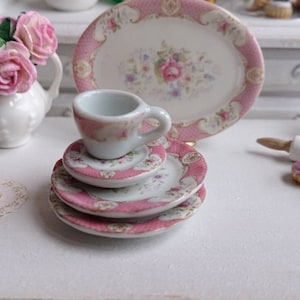 May include: Miniature porcelain tea set with pink floral and gold trim details. The set includes a teacup, saucers, and a serving plate. A small white vase with pink roses and a miniature cake are also visible.