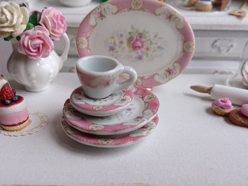 May include: Miniature porcelain tea set with pink floral and gold trim details. The set includes a teacup, saucers, and a serving plate. A small white vase with pink roses and a miniature cake are also visible.