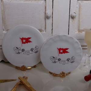 Könnte beinhalten: Zwei weiße Keramikteller mit einem roten und weißen White Star Line Flaggen-Design. Die Teller stehen auf goldenen Metallständern. Der Text auf den Tellern lautet "White Star Line 1848".