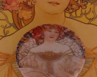 May include: A round, glass cabochon featuring a vintage illustration of a woman in a white dress with a floral crown. The woman is surrounded by a decorative, Art Nouveau-style frame.