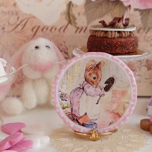 May include: A pink and white ceramic plate with a picture of a mouse holding a mirror. The plate is on a gold stand and there are pink and white heart-shaped candies in the background.