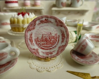 May include: A small, pink and white porcelain plate with a red and white floral design and a picture of a bridge. The plate is on a white doily.