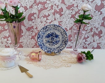 Op de afbeelding: Een blauw en wit porseleinen bord met een bloemenmotief staat op een gouden standaard. Het bord is omgeven door een witte kantonderlegger. Twee roze glazen vazen met roze rozen staan aan weerszijden van het bord. Een roze glazen kelk en een witte cupcake met roze glazuur bevinden zich op de voorgrond.