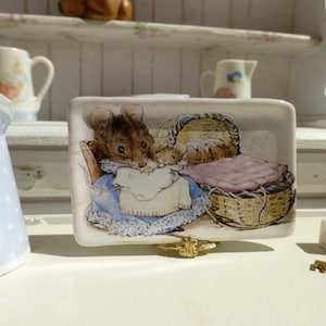 Puede incluir: Un plato de cerámica blanco con una imagen de un ratón marrón sosteniendo un ratón bebé en una cesta de mimbre con una manta rosa. El plato está sobre un soporte dorado.