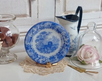 May include: A small blue and white porcelain plate with a gold stand. The plate features a blue and white floral design with a scene of a deer in a forest. The plate is on a white lace doily.