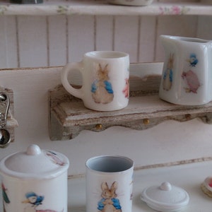 Op de afbeelding: Een set miniatuur witte keramische bekers en een kan met een fantasierijk ontwerp met Peter Rabbit en andere personages. De bekers en de kan worden getoond op een klein houten plankje met een gegolfde rand.