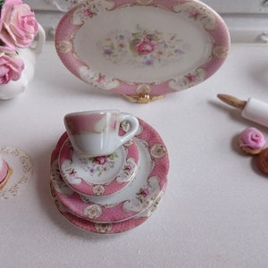 May include: Miniature porcelain tea set with pink floral accents, including a teacup, saucer, and plates. A small cake and decorative flowers are also present, creating a charming, whimsical scene.