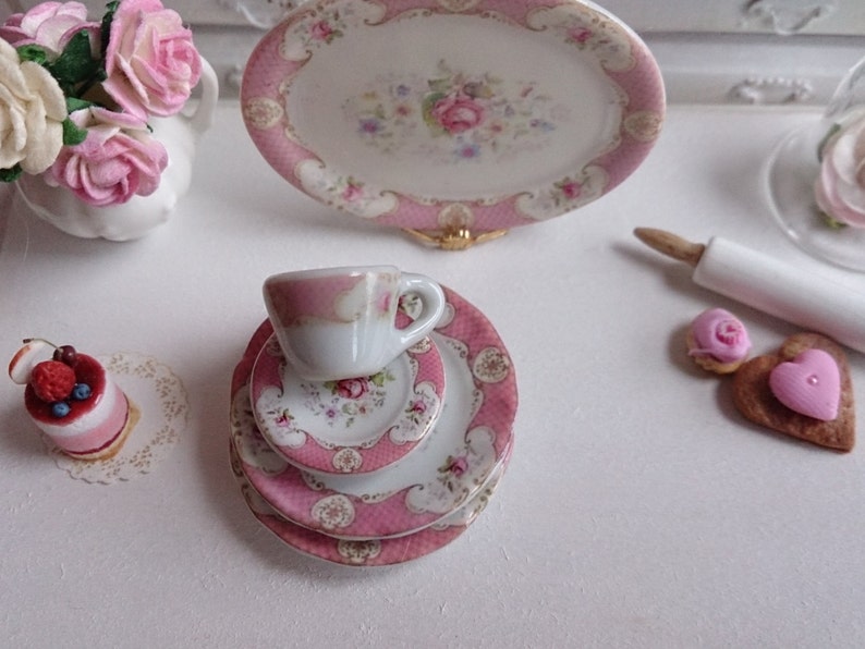 May include: Miniature porcelain tea set with pink floral accents, including a teacup, saucer, and plates. A small cake and decorative flowers are also present, creating a charming, whimsical scene.