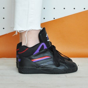 May include: Black high-top sneakers with purple and red accents. The sneakers have a lace-up closure and a padded collar. The shoes are worn with white jeans with a frayed hem. The background is white with black dots and orange.