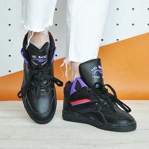 May include: Black high-top sneakers with purple and red accents. The sneakers have the words "VIG BANG LIGHT SHOE" printed on the side. The shoes are paired with white, distressed jeans.