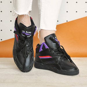 May include: Black high-top sneakers with purple and red accents. The sneakers have the words "Vig Bang Light Shoe" printed on the tongue. The shoes are worn with white jeans with frayed hems.