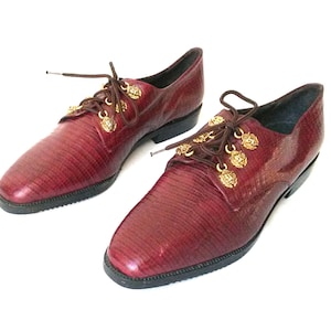 May include: Burgundy leather dress shoes with a textured finish. The shoes feature brown laces and gold-coloured decorative accents. The soles are black, and the shoes are set against a white background.