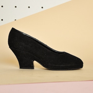 May include: A single black suede pump with a rounded toe and a unique, curved heel. The shoe is set against a backdrop of beige and white geometric shapes, highlighting its classic design.