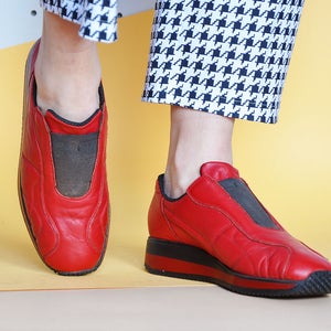 May include: Red leather slip-on sneakers with a black sole and a black elastic insert. The shoes have a quilted design and are shown against a yellow and white background. The shoes are a bold fashion statement.