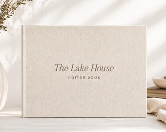 May include: A rectangular guest book with a light beige fabric cover. The front cover features the centered text "The Lake House" in a dark serif font, with the words "VISITOR BOOK" printed in a smaller, simpler font directly underneath.