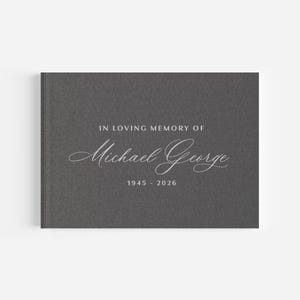 May include: A charcoal grey landscape-oriented book with a textured fabric cover. The front cover features white text that reads IN LOVING MEMORY OF Michael George 1945 - 2026, with the name Michael George written in a decorative script font.