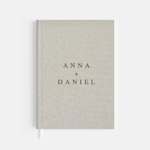 May include: A light gray linen-covered journal with the names "ANNA + DANIEL" printed in dark brown. The journal has a matching ribbon bookmark. The cover has a textured appearance.