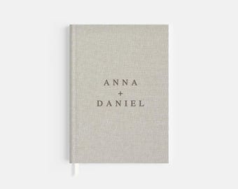 May include: A light gray linen-covered journal with the names "ANNA + DANIEL" printed in dark brown. The journal has a matching ribbon bookmark. The cover has a textured appearance.