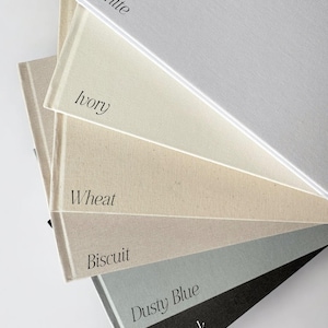 May include: A stack of six fabric-covered books arranged in a fan shape. The covers are labelled with the text White, Ivory, Wheat, Biscuit, Dusty Blue, and Black, each corresponding to the specific colour of the textured material on the book cover.