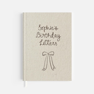 Może przedstawiać: Beżowy notatnik w płóciennej okładce z napisem "Sophie's Birthday Letters" brązową czcionką. Pod tekstem znajduje się brązowa ilustracja kokardy. Notatnik ma białą wstążkę jako zakładkę.