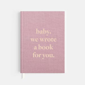 May include: A pink linen-covered journal with the text "baby, we wrote a book for you" in beige. The journal has a white ribbon bookmark. The cover has a textured appearance.