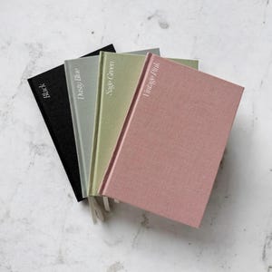 Puede incluir: Cuatro diarios de tapa dura en negro, azul polvoriento, verde salvia y rosa vintage. Cada diario tiene una cinta de marcador a juego y el nombre del color impreso en blanco en la portada. Los diarios están dispuestos en forma de abanico sobre una superficie de mármol.