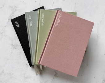 May include: Four hardcover journals in black, dusty blue, sage green, and vintage pink. Each journal has a matching ribbon bookmark and the color name printed on the cover in white. The journals are arranged in a fan shape on a marble surface.