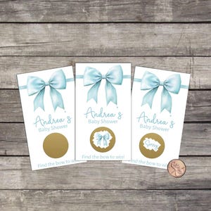 May include: Three baby shower scratch-off cards with a light blue bow design. The cards read "Andrea's Baby Shower" and "Find the bow to win!". One card has a gold scratch-off area, another has a bow image, and the last says "Sorry".