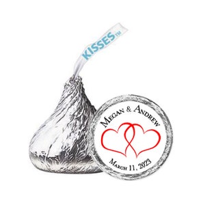 May include: White round sticker with black text that reads "Megan & Andrew" and "March 11, 2023". Two red intertwined hearts are in the center of the sticker. The sticker is on top of a silver Hershey's Kisses chocolate.
