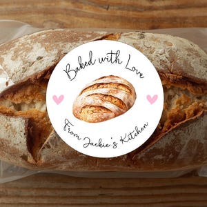May include: A loaf of freshly baked bread in a clear plastic bag. A round sticker on the bag reads "Baked with Love From Jackie's Kitchen" with a watercolor illustration of a loaf of bread and two pink hearts.