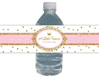 May include: Water bottle label with a pink and gold glitter design. The label features a gold crown and the text "A Little Princess Is On Her Way".