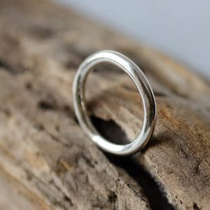 Può includere: Un anello d'argento dal design liscio e arrotondato. L'anello è appoggiato su una superficie di legno strutturata, marrone chiaro. La superficie lucida dell'anello riflette la luce, evidenziando la sua forma circolare.