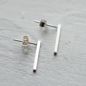 Peut inclure: Une paire de boucles d'oreilles minimalistes en argent en forme de barres. Les boucles d'oreilles sont de simples barres droites avec un fermoir et un dos. Elles sont présentées sur une surface gris clair.