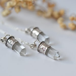 Puede incluir: Tres collares de cristal de cuarzo transparente con detalles metálicos plateados. Los cristales son cilíndricos con extremos puntiagudos. Los collares están sobre una superficie blanca con un fondo borroso de flores secas.